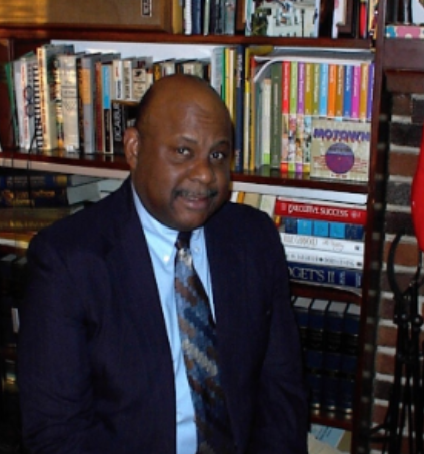 Photo of Jack Cooper. He is a black man wearing a suit and tie, standing in front of a bookcase.