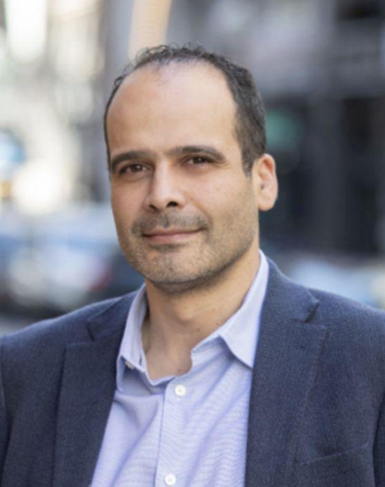 A smiling man in a suit in front of a blurred city background