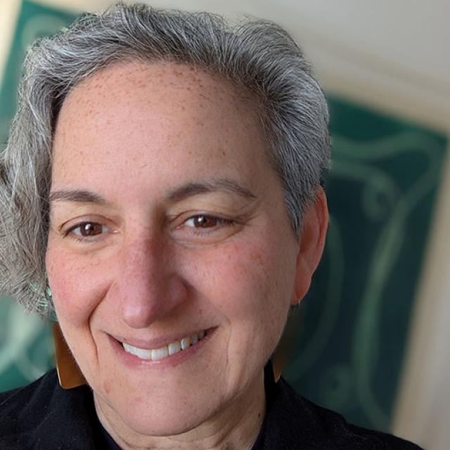 Photo of Jennifer, a white woman with gray hair. She is smiling at the camera. The background is out of focus but shows a blue green art piece. 