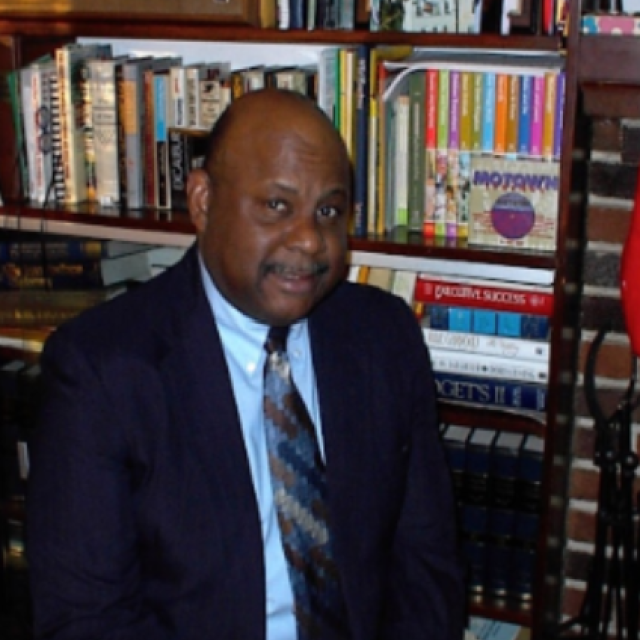 Photo of Jack Cooper. He is a black man wearing a suit and tie, standing in front of a bookcase.