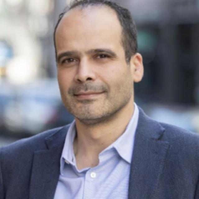 A smiling man in a suit in front of a blurred city background